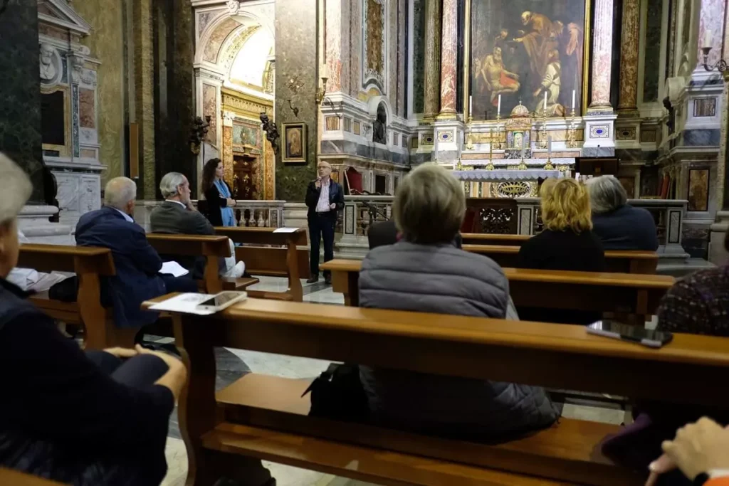 Interno di chiesa con persone fotografate di spalle e sul fondo una persona che parla al microfono