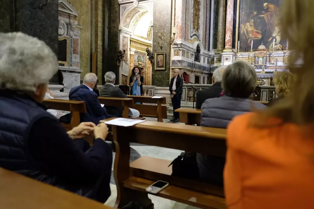 Interno di chiesa con persone fotografate di spalle e sul fondo una persona che parla al microfono