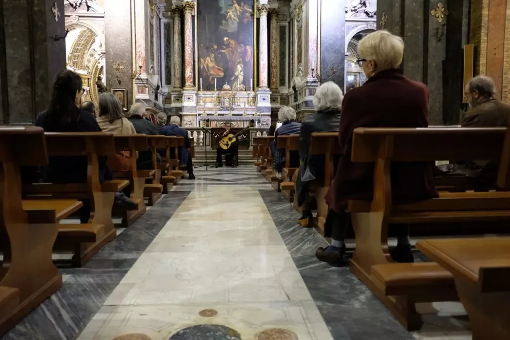 Fotografia della navata centrale di una chiesa con uditori seduti e una persona seduto sui gradini del sagrato con una chitarra